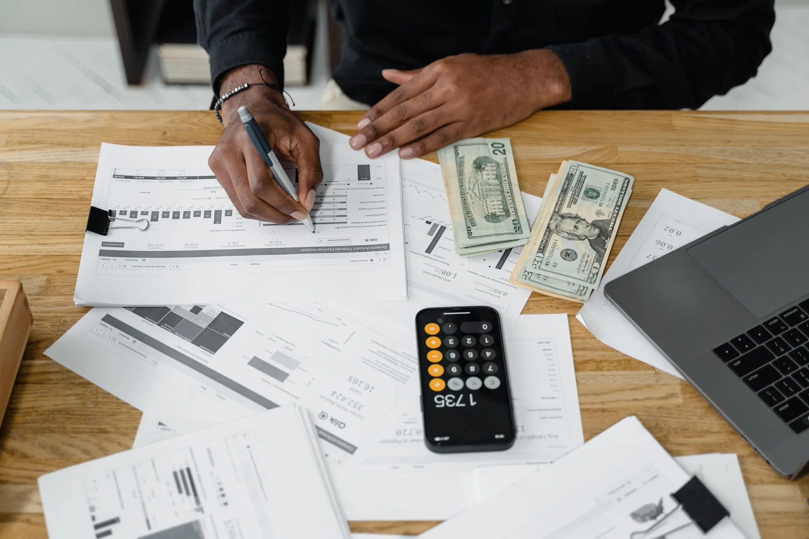 Pessoa revisando papéis com calculadora, notas de dólar e laptop sobre a mesa