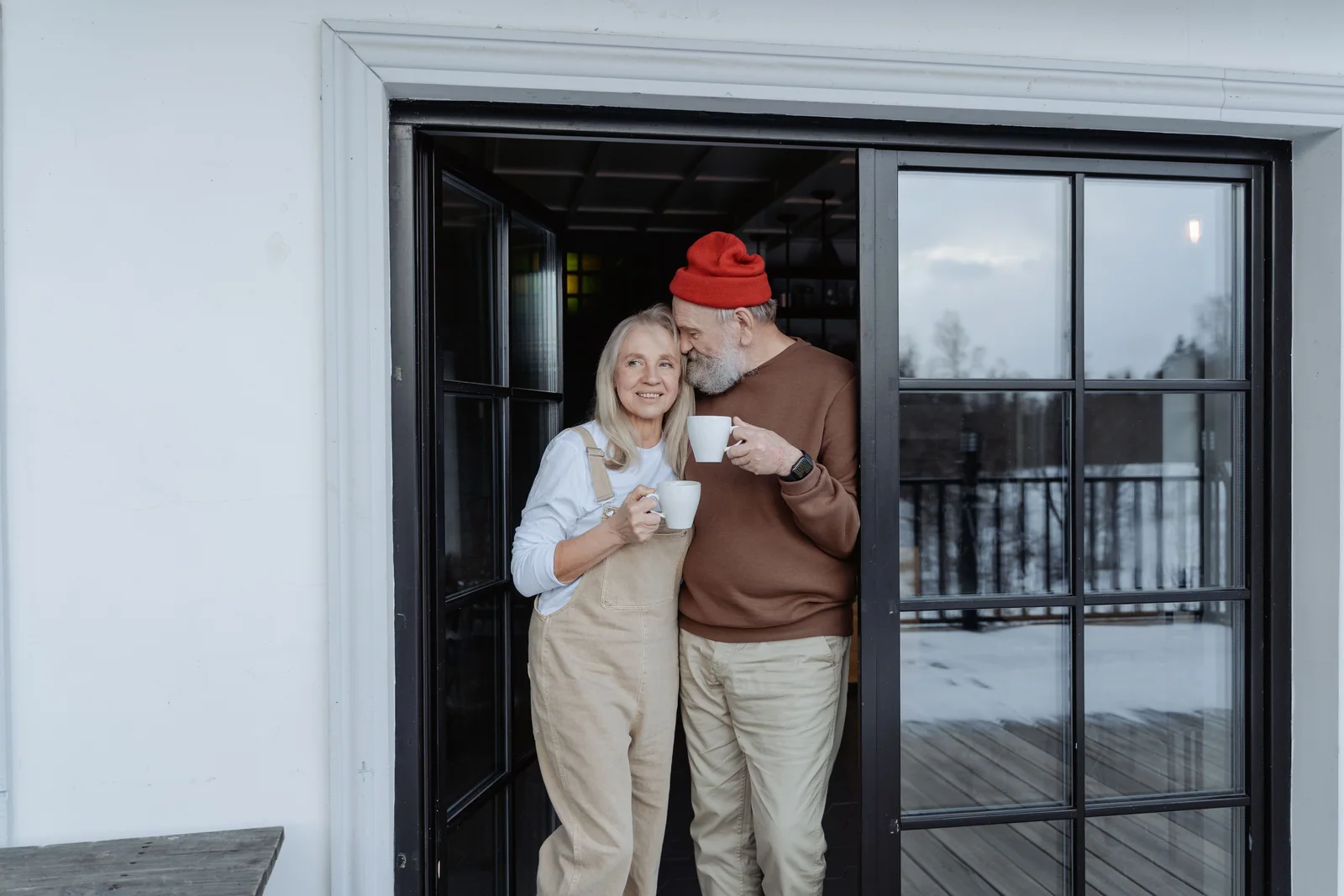 Casal mais velho na porta de casa, cada um com uma xícara de café