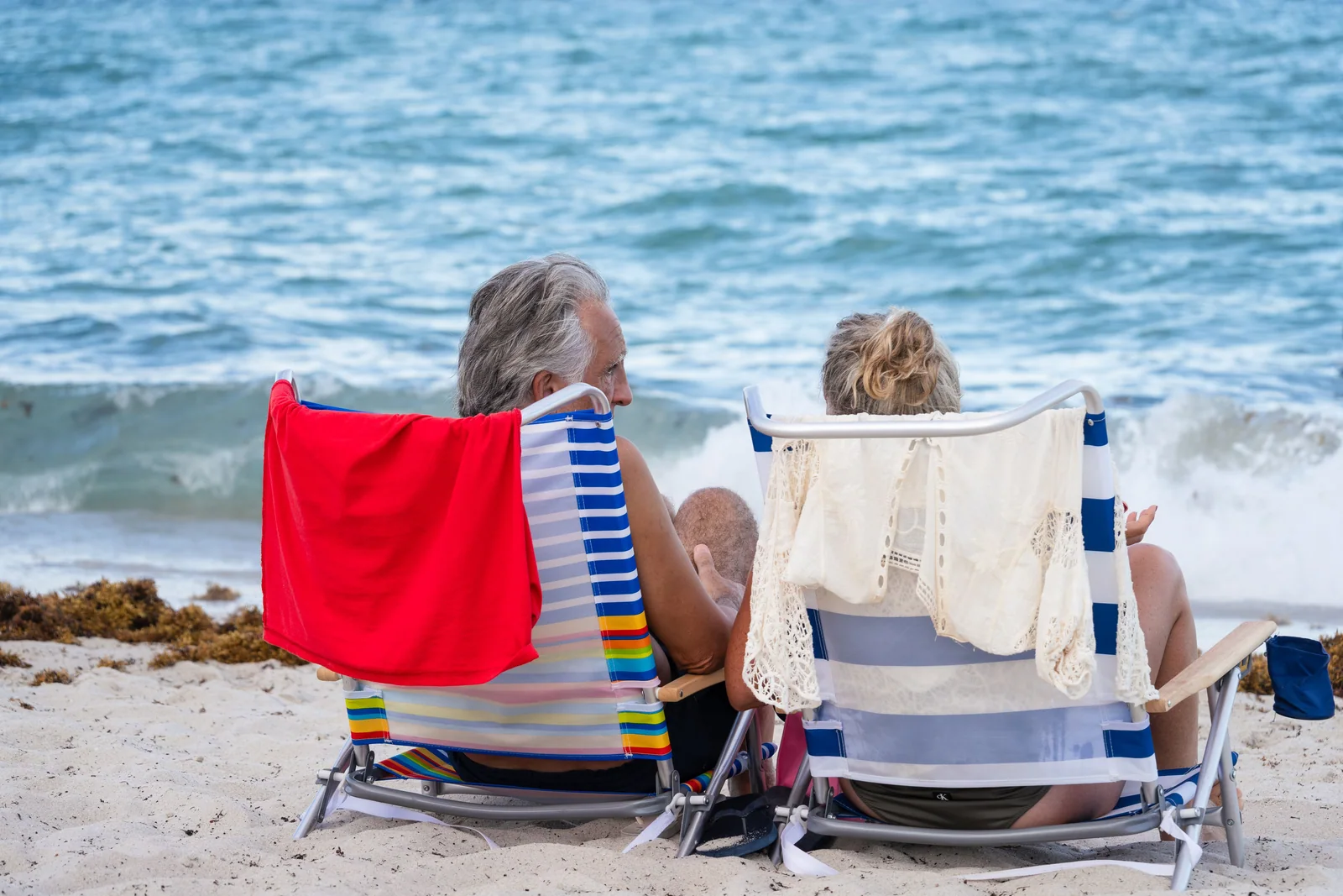 Casal sentado de costas em cadeiras de praia, olhando o mar