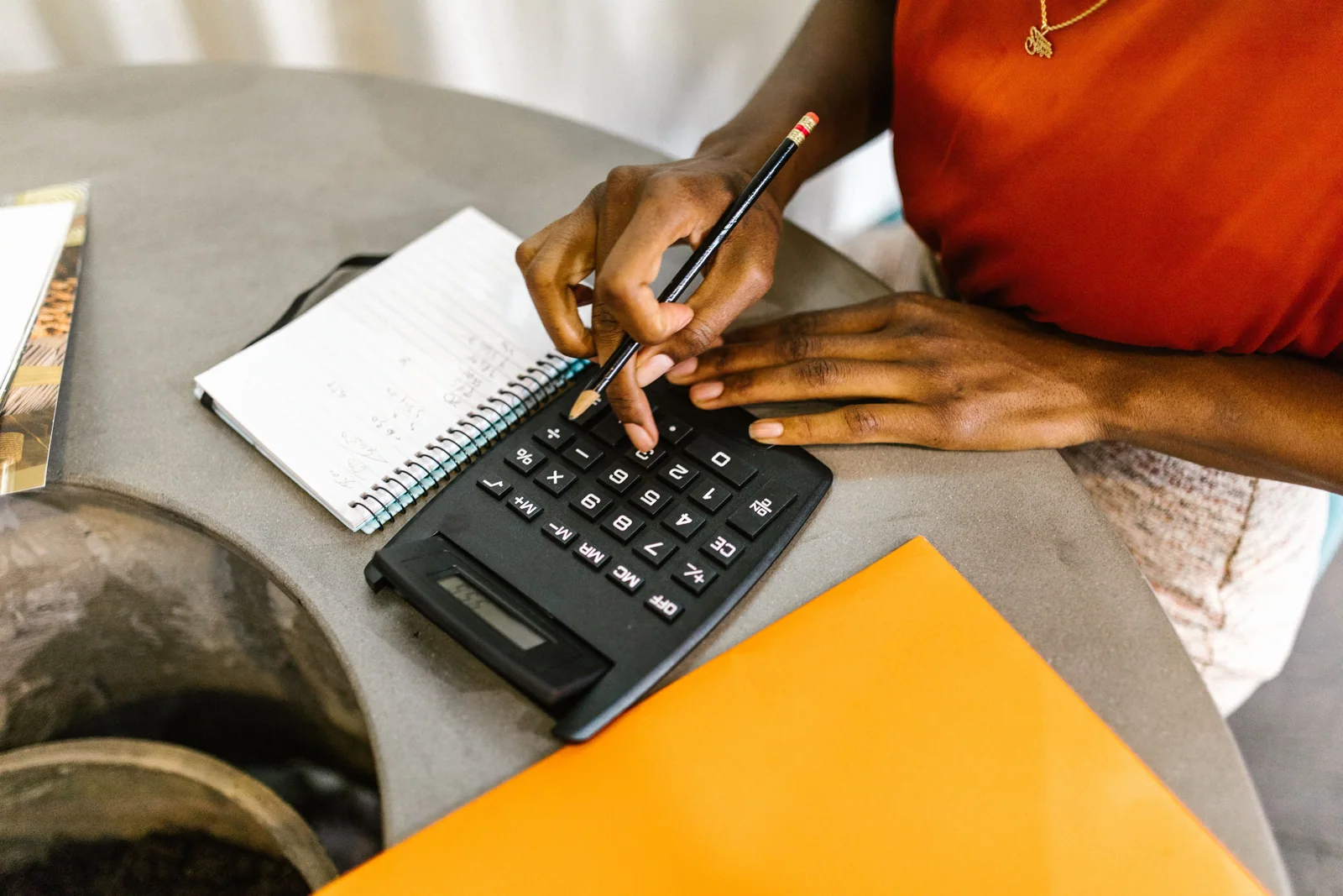 Mãos digitando em uma calculadora preta com um lápis, ao lado de um caderno de anotações e de uma pasta laranja