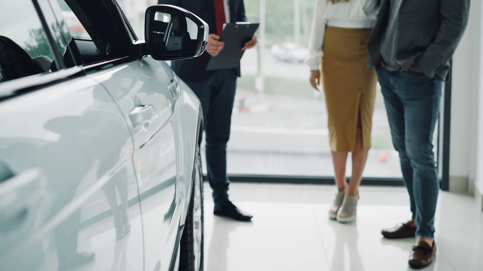 Carro claro em primeiro plano numa concessionária, com compradores e vendedor ao fundo